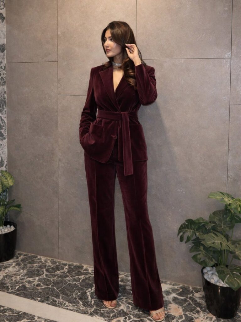 Woman wearing a burgundy velvet blazer and matching wide-leg trousers against a grey wall.