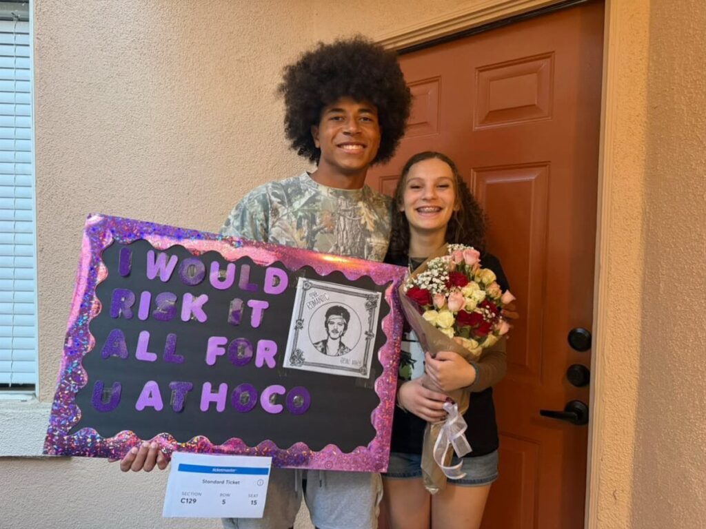 Teenage boy holding a Bruno Mars themed homecoming proposal poster and a girl with flowers.