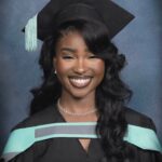 Woman with wavy black hair wearing a graduation cap and gown smiling.