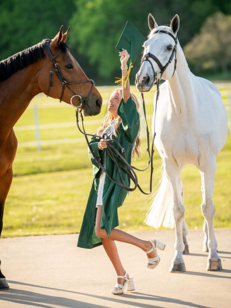 Graduate in green cap and gown posing between a brown and white horse.