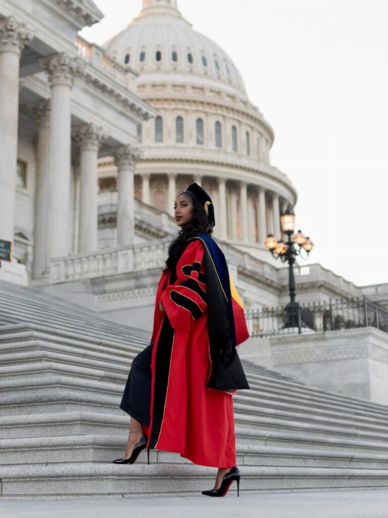 Woman in red doctoral graduation gown and cap posing on Capitol building steps.