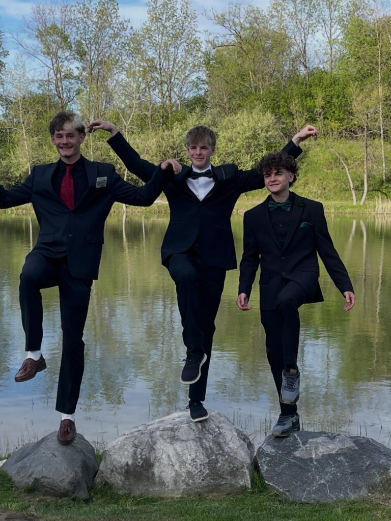 Three boys in black suits posing playfully on rocks by a pond.