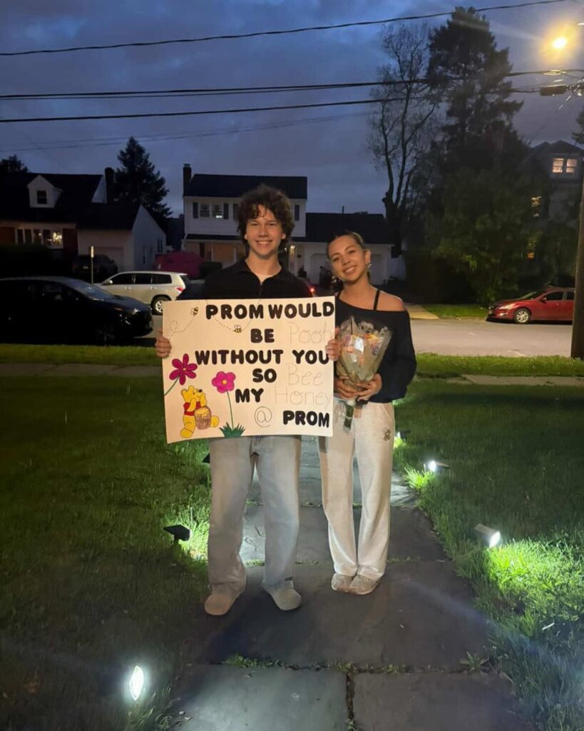 Teenage couple standing on a walkway with a prom proposal poster and flowers.