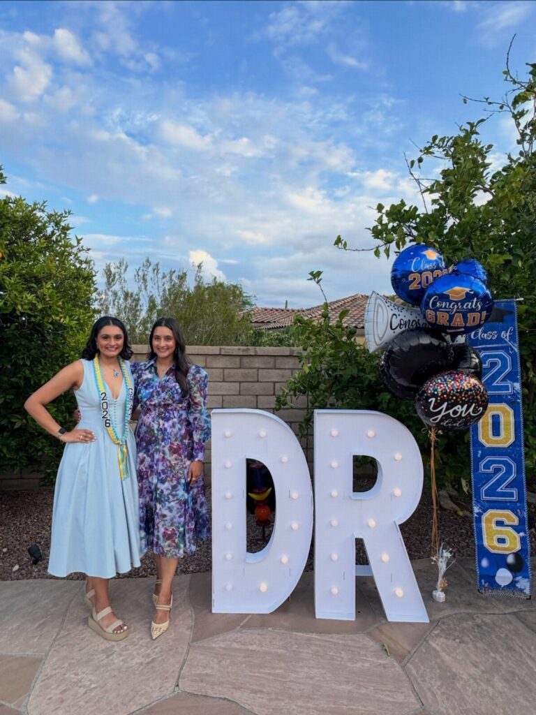 Two women in dresses posing by large lit DR letters and graduation balloons.