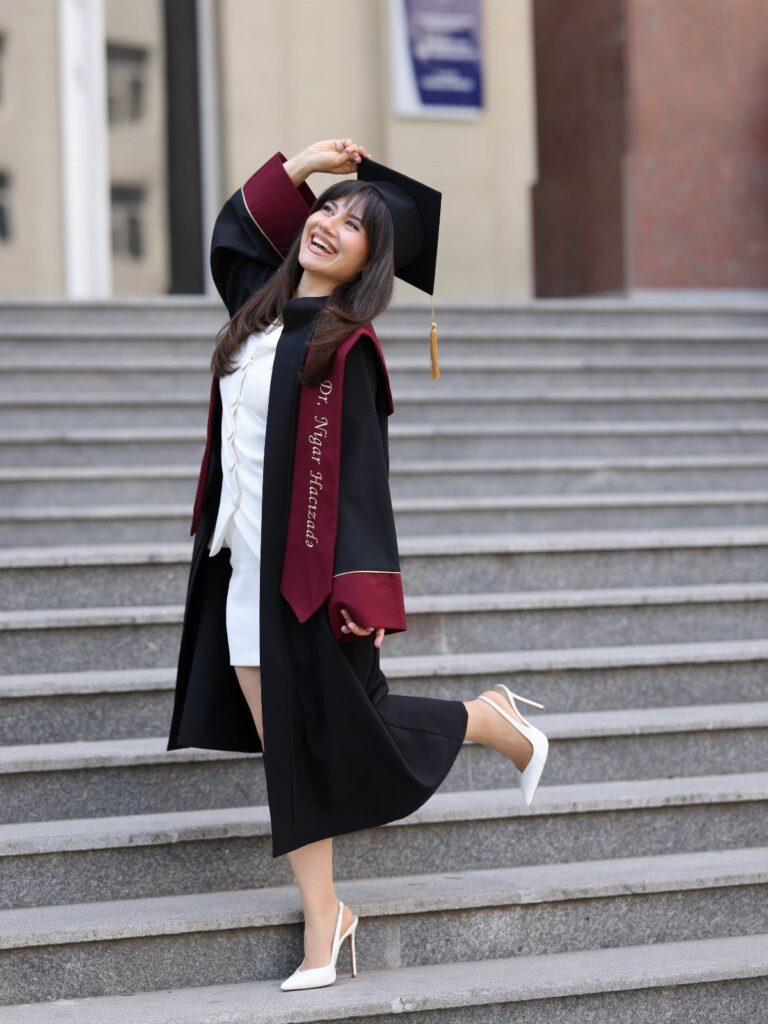 Woman in graduation gown and cap posing on stone stairs with a smile.