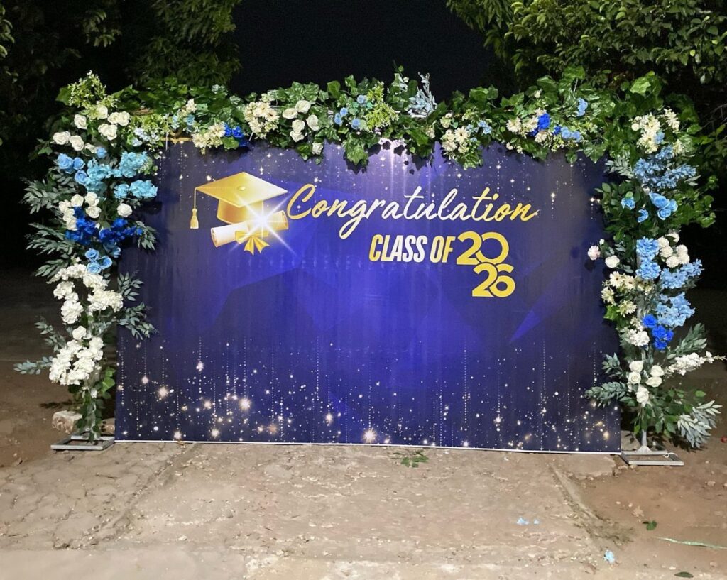 Blue graduation backdrop with gold lettering framed by white and blue floral arrangements.