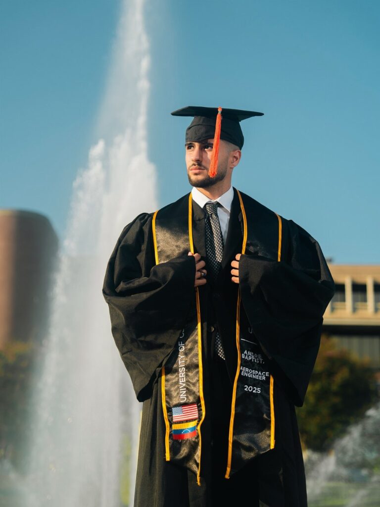 Graduate in black cap and gown posing in front of a large water fountain