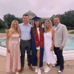 Graduating woman in blue gown posing with four family members by a campus fountain.