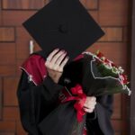 Graduate in black gown holding a mortarboard over their face and red roses.