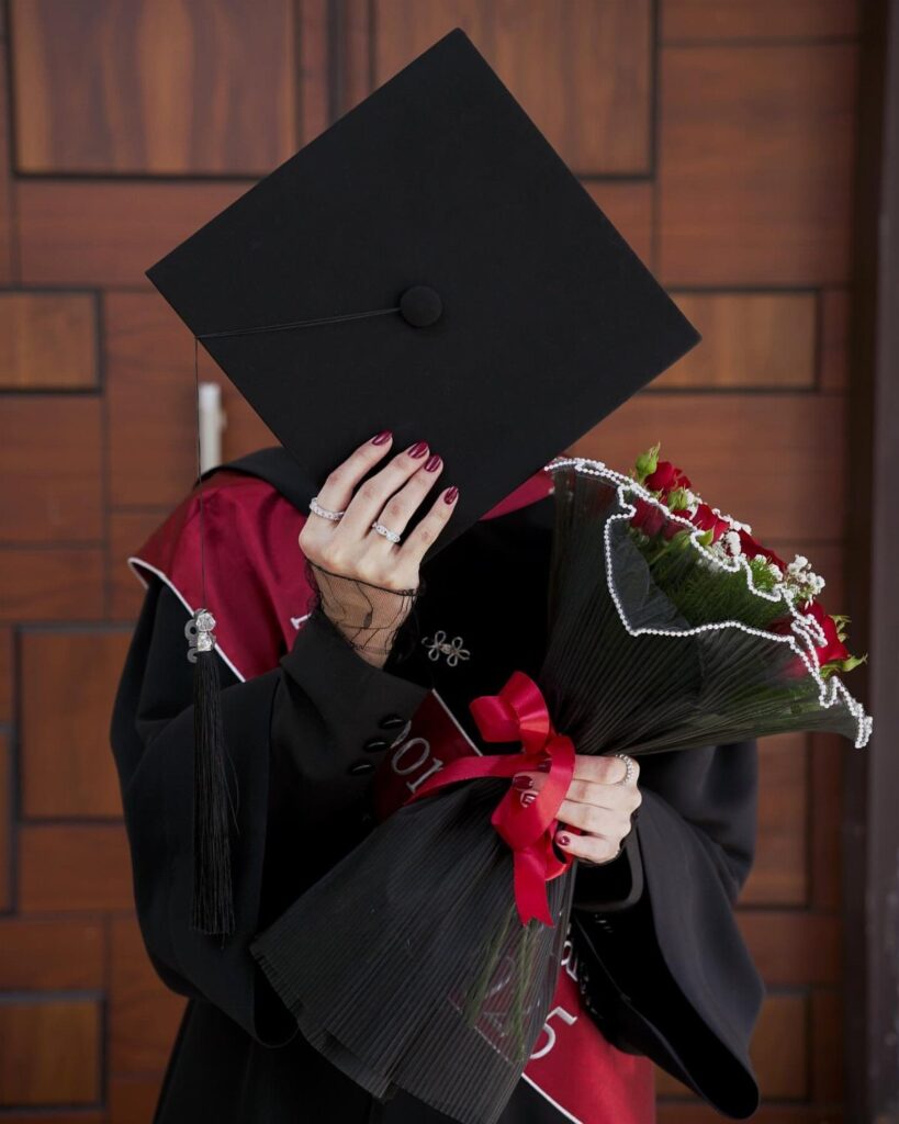Graduate in black gown holding a mortarboard over their face and red roses.