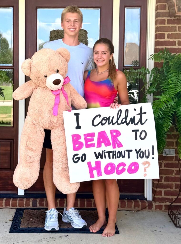 Teenage couple posing with a giant teddy bear and a homecoming proposal sign.