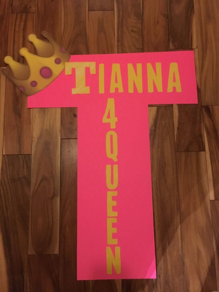 Pink T-shaped poster board with yellow text and a crown on wooden floor