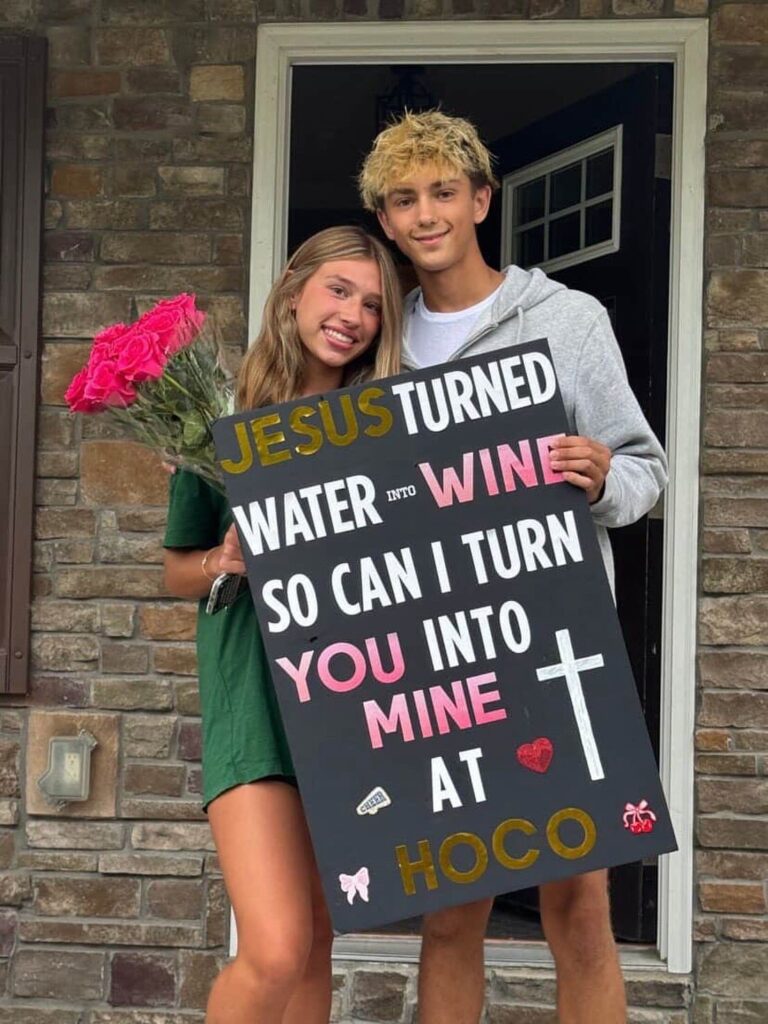 Teenage couple posing with a homecoming proposal sign and pink roses by a door