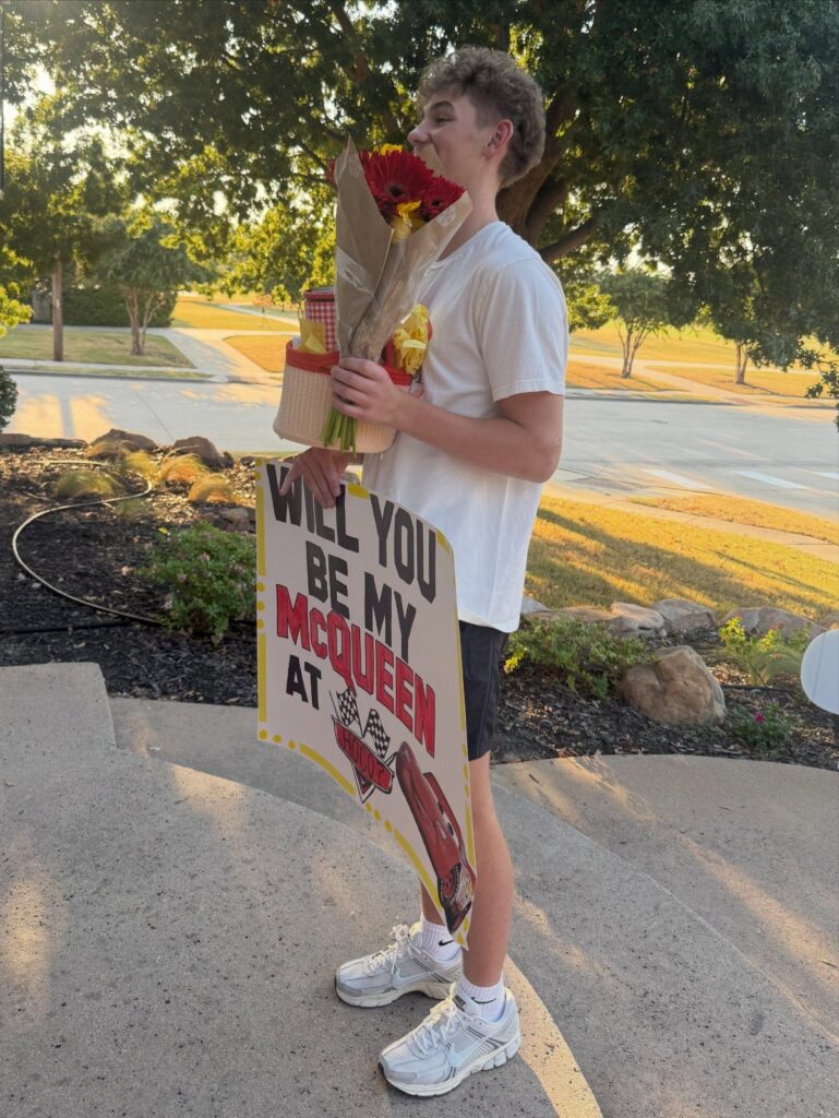 Teenage boy holding flowers and a Lightning McQueen themed promposal sign outdoors.