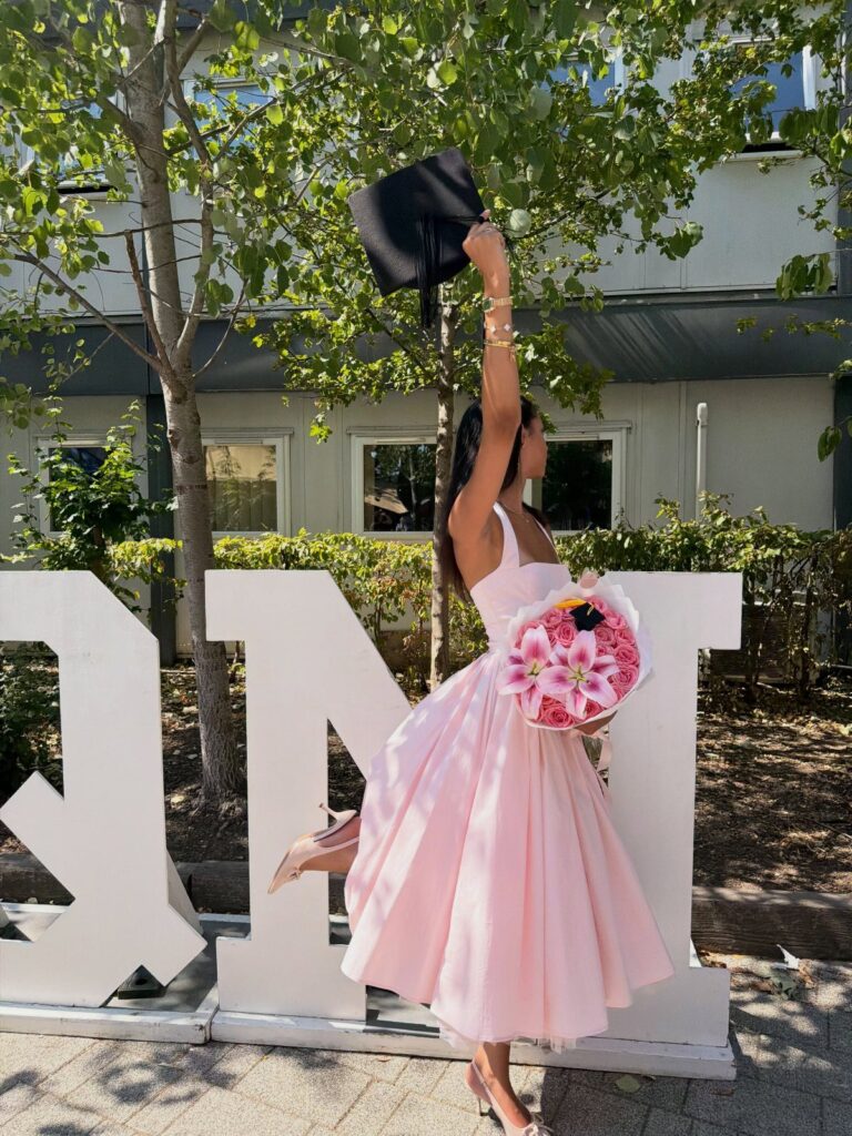 Woman in a pink midi dress holding a graduation cap and flower bouquet.