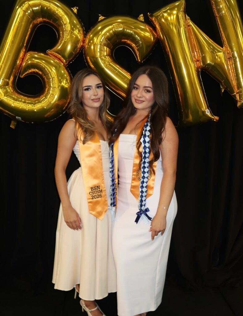 Two women in white dresses and gold sashes posing before gold balloons.
