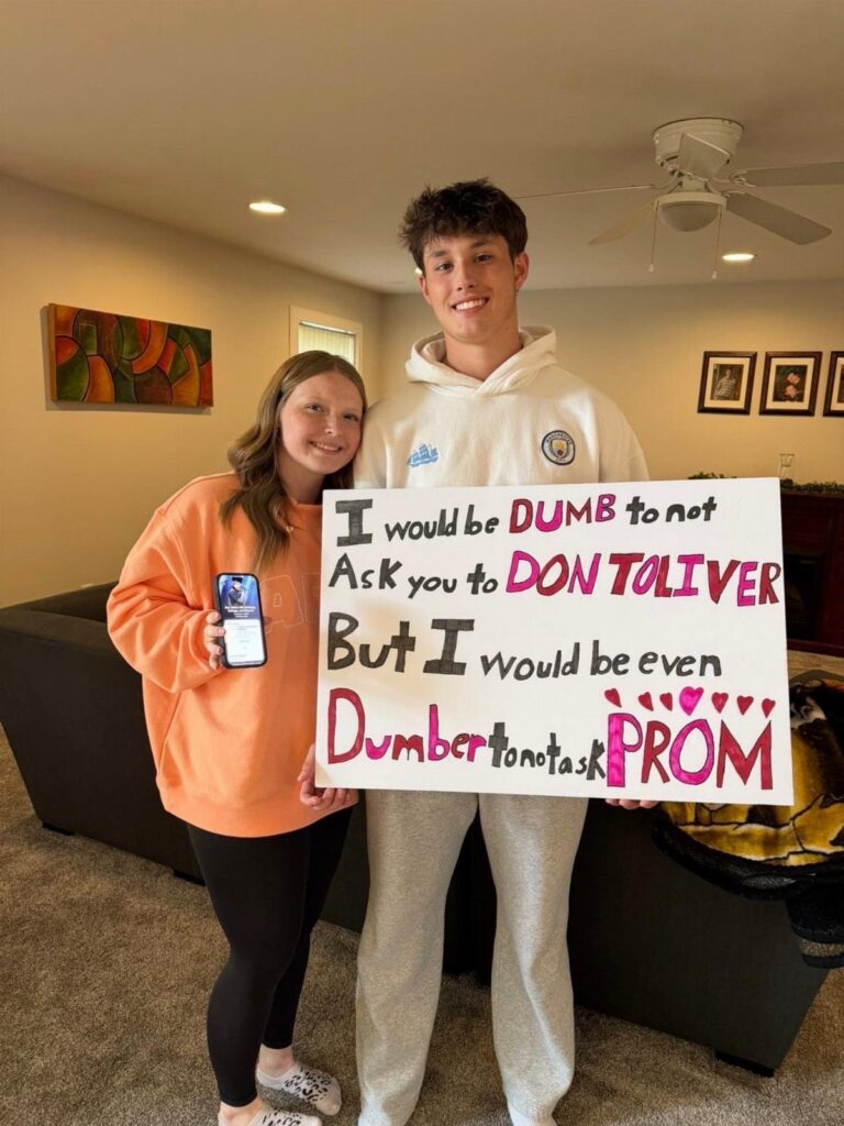 Teenage couple posing with a handmade promposal poster in a living room
