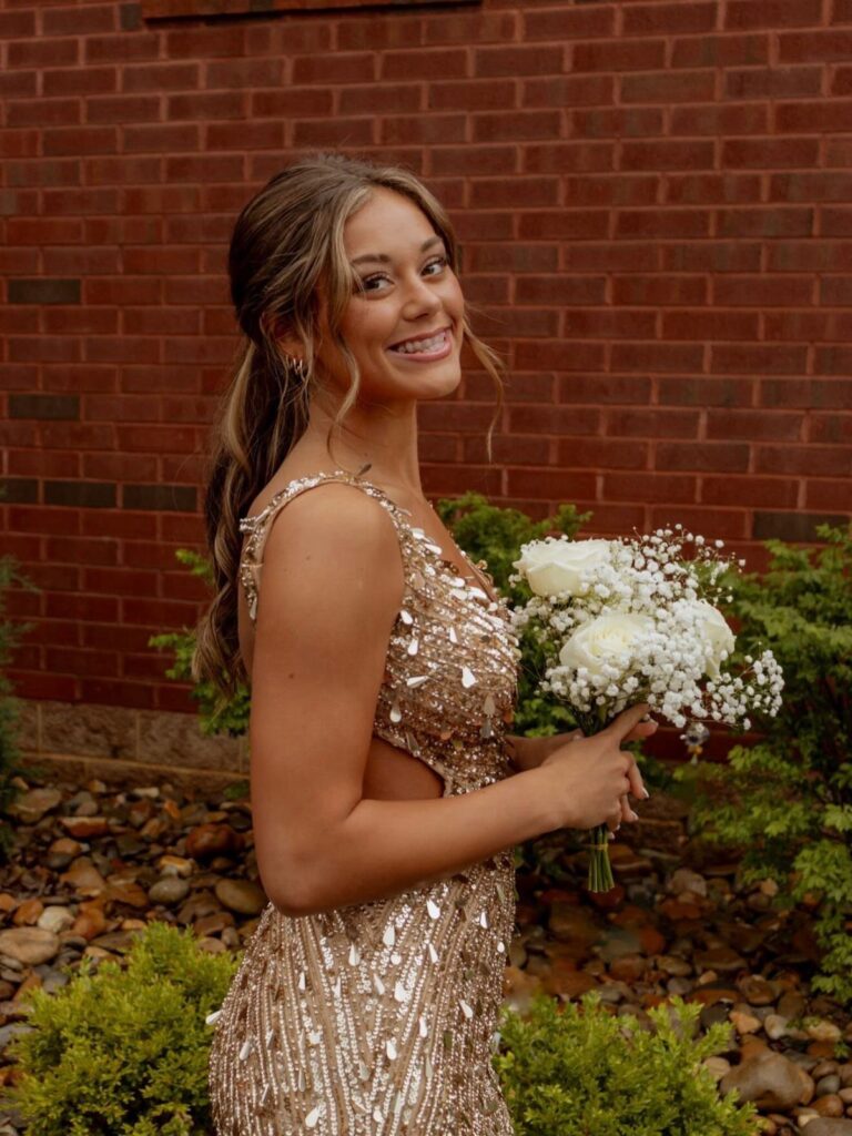 Young woman in gold sequined dress glancing over shoulder holding white flowers.