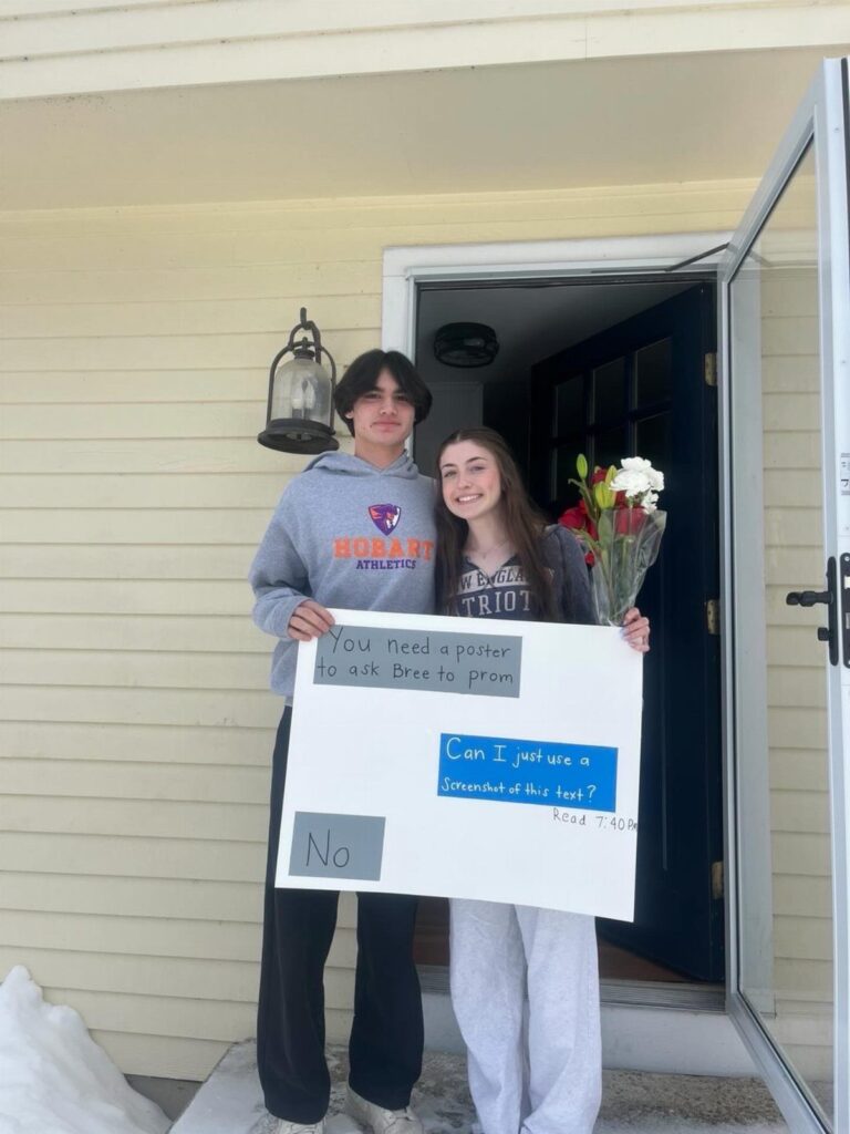 Teenage boy and girl holding a prom proposal poster and flowers in a doorway