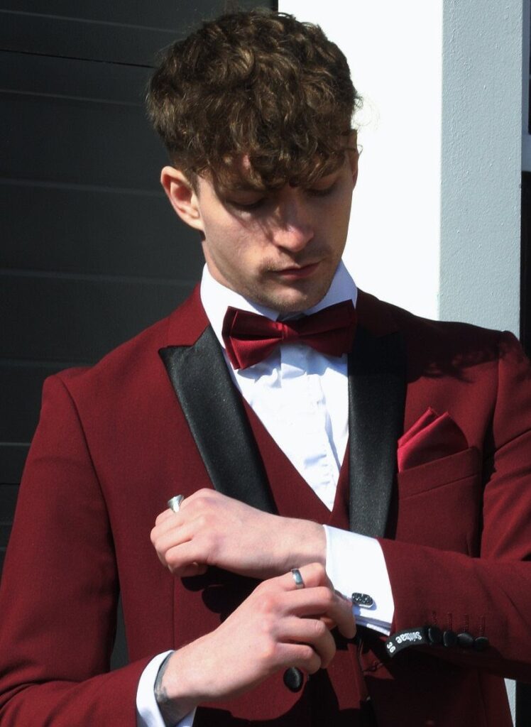 Young man in a burgundy tuxedo with black lapels adjusting his cufflink.