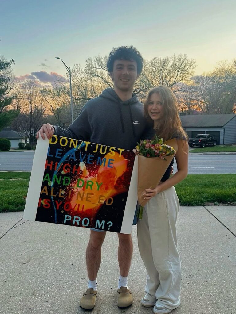 Teenage couple posing outdoors with a promposal poster and a bouquet of flowers.