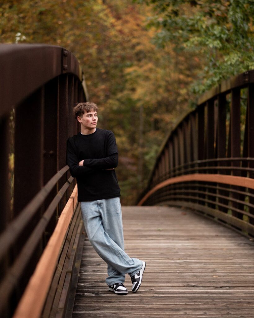 Young man posing on a wooden bridge surrounded by autumn foliage for senior portraits.