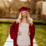 Young woman in red graduation cap and gown posing on a stone path.