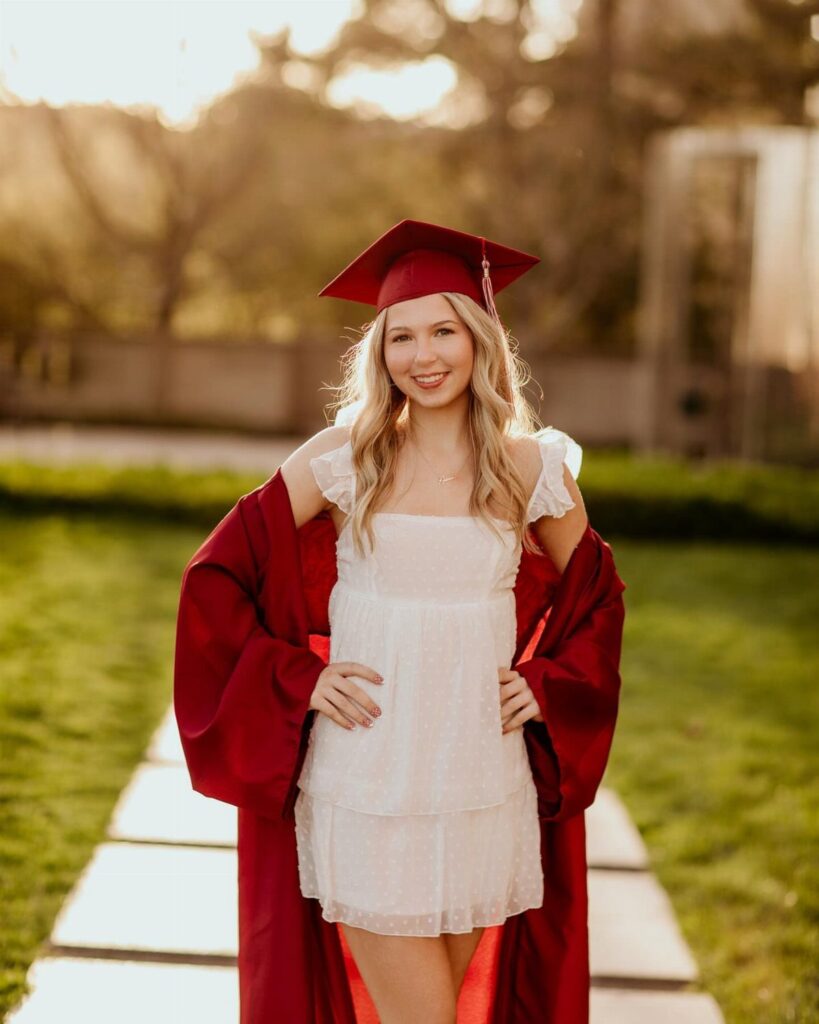 Young woman in red graduation cap and gown posing on a stone path.