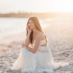 Young woman in a long white dress sitting on a sandy beach at sunset.