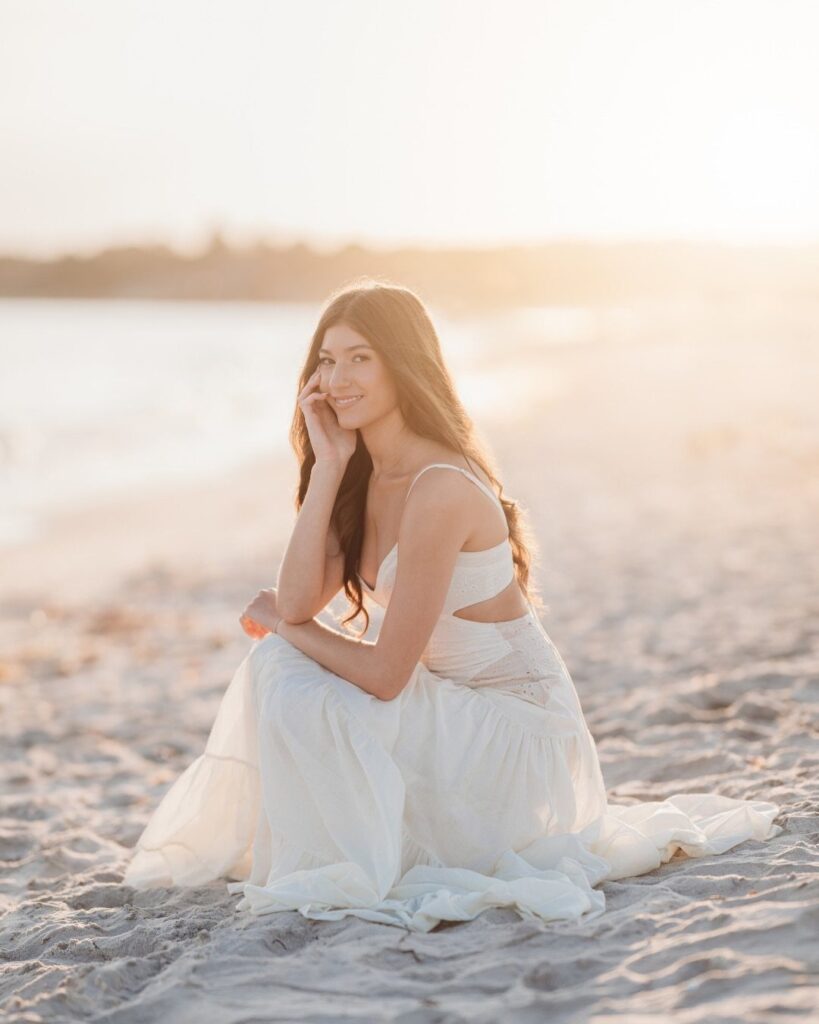Young woman in a long white dress sitting on a sandy beach at sunset.