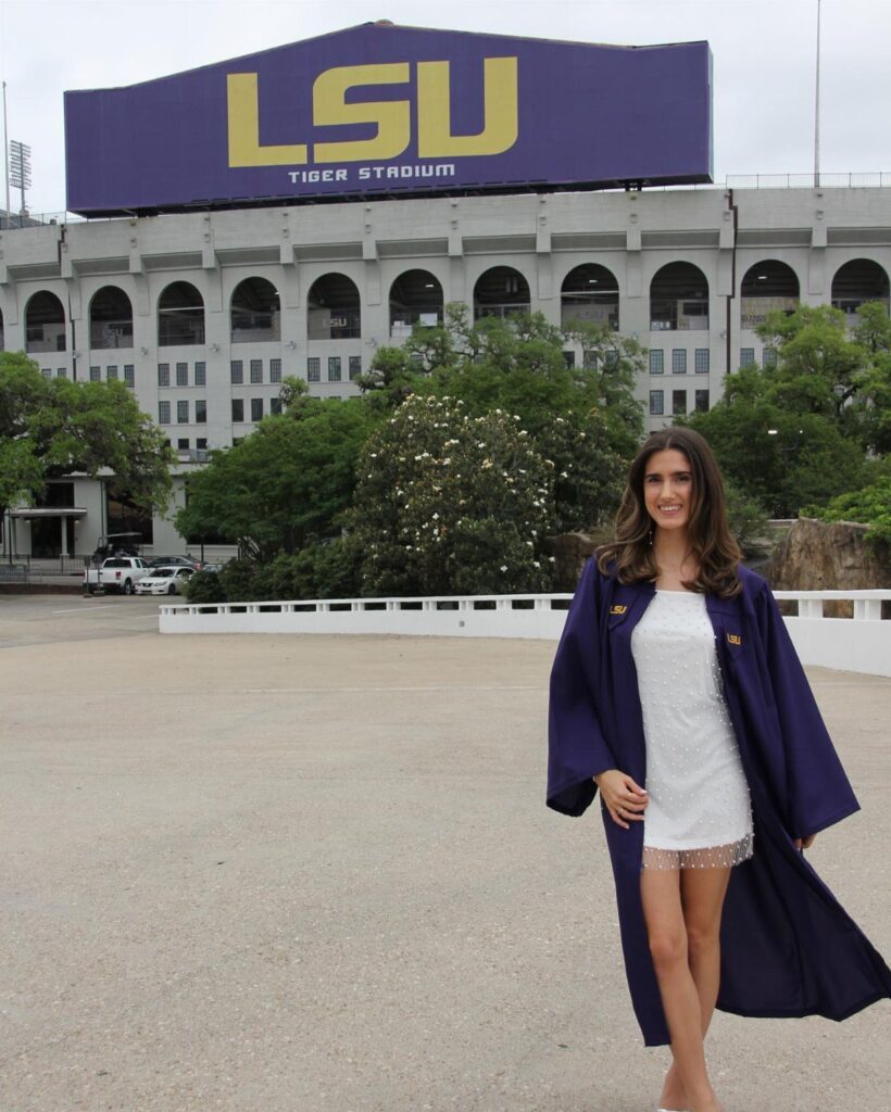 Young woman in a white dress and purple graduation gown before LSU stadium.