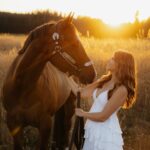 Young woman in white dress posing with a brown horse in a sunset field.