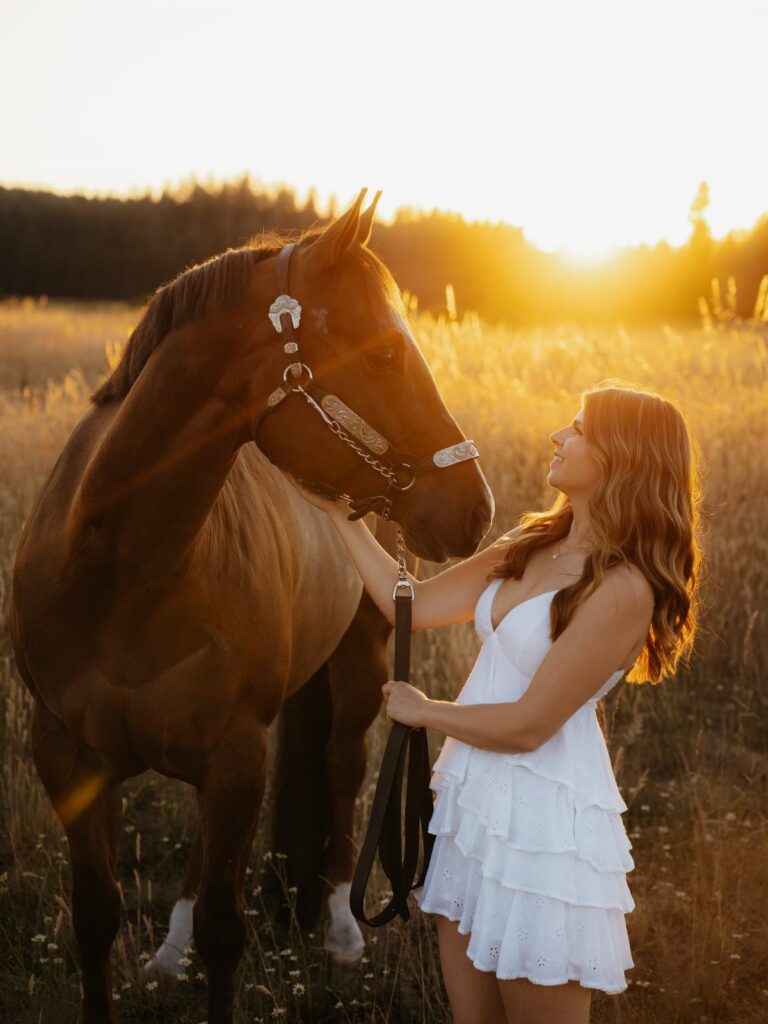 Young woman in white dress posing with a brown horse in a sunset field.