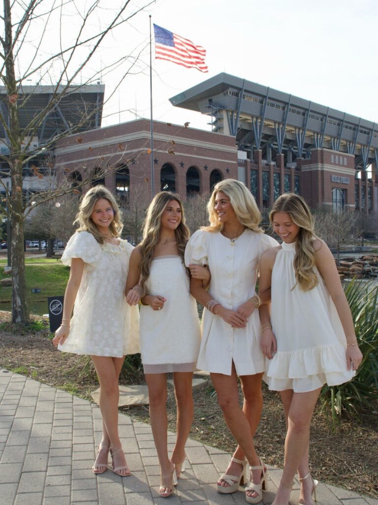 Four women in short white dresses and heels posing in front of a stadium.