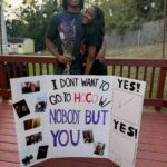 Teenage couple posing behind a handmade homecoming proposal poster on a wooden deck