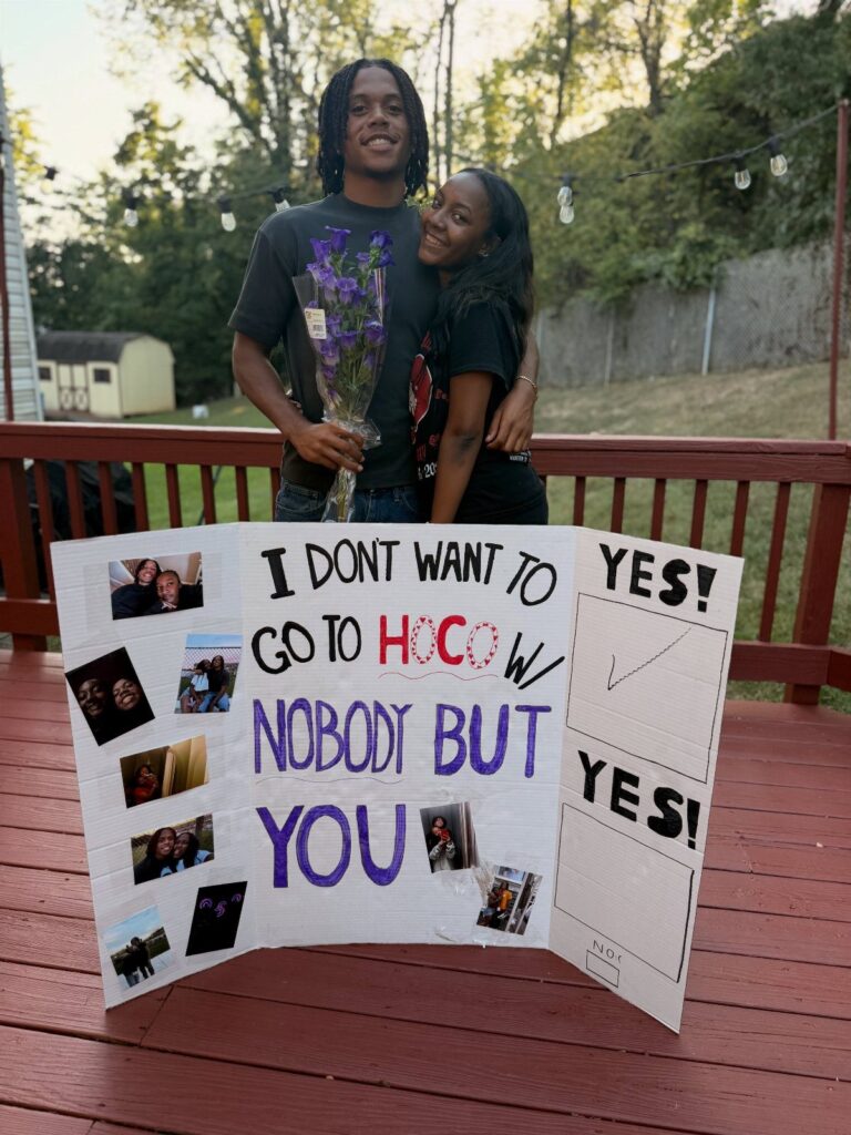 Teenage couple posing behind a handmade homecoming proposal poster on a wooden deck